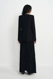 A model shown from the back wearing a black long-sleeve blazer and a long black pleated skirt, standing against a plain white studio background.