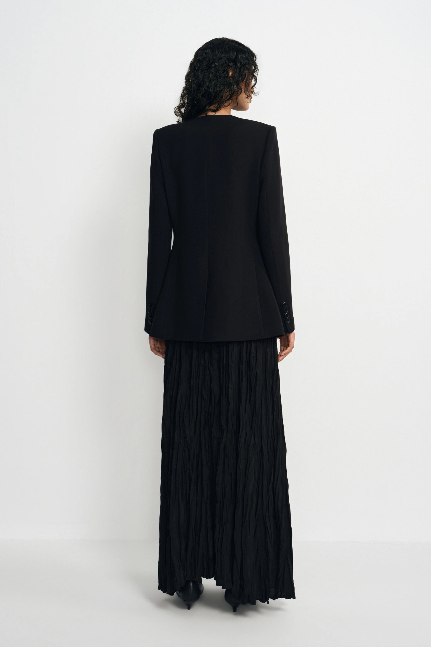 A model shown from the back wearing a black long-sleeve blazer and a long black pleated skirt, standing against a plain white studio background.
