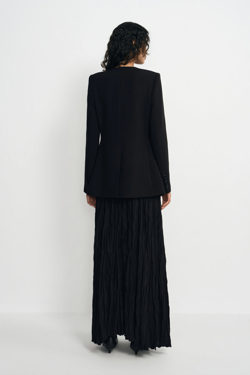 A model shown from the back wearing a black long-sleeve blazer and a long black pleated skirt, standing against a plain white studio background.