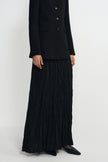A model wearing a black double-breasted blazer and a long, black pleated maxi skirt, paired with black pointed-toe flats, photographed against a plain light gray studio background.