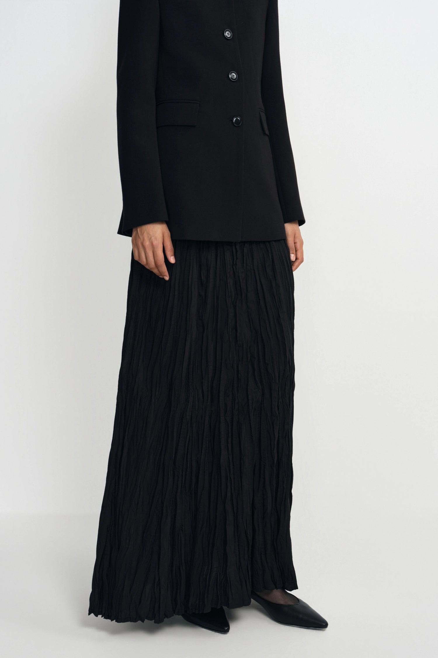 A model wearing a black double-breasted blazer and a long, black pleated maxi skirt, paired with black pointed-toe flats, photographed against a plain light gray studio background.