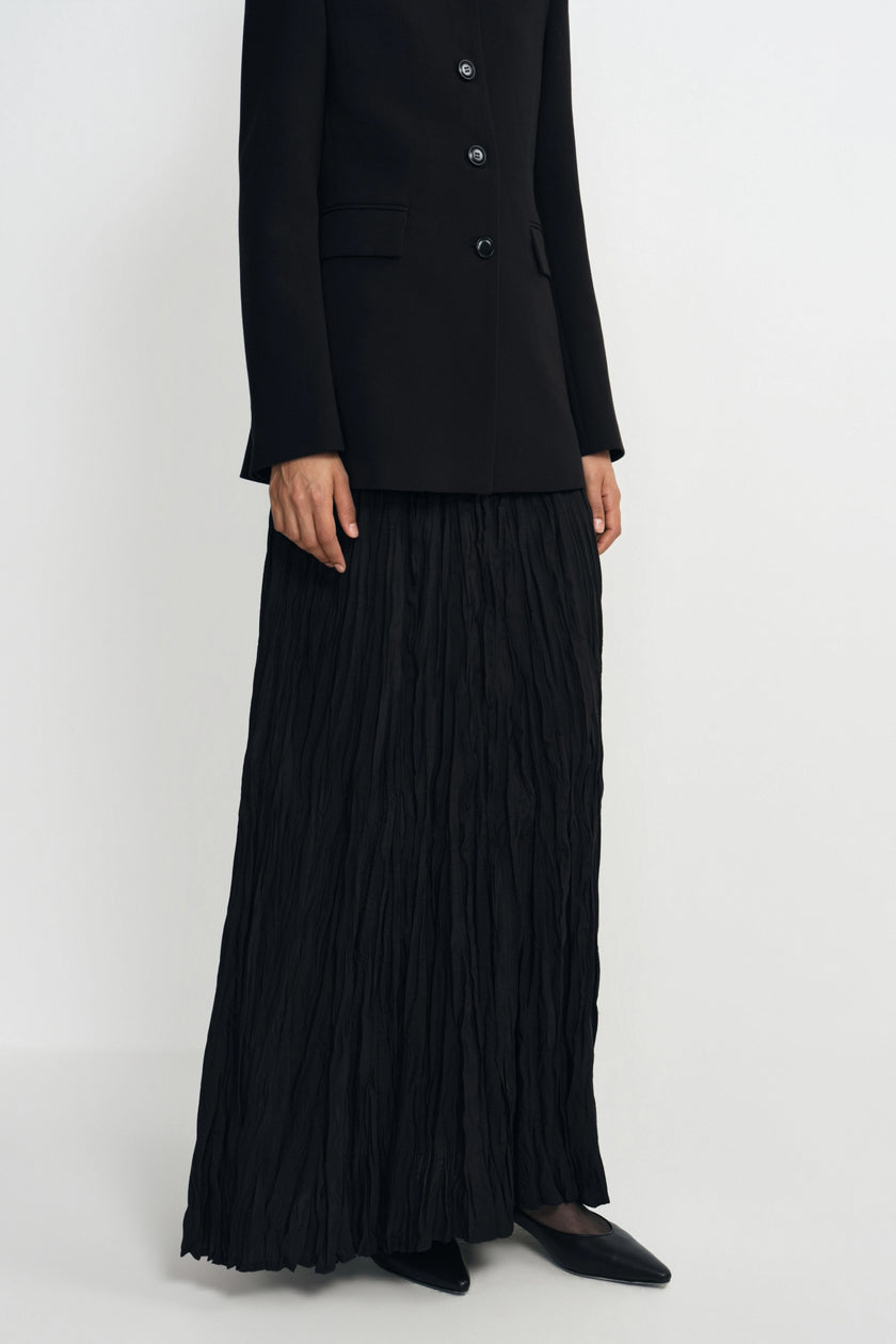 A model wearing a black double-breasted blazer and a long, black pleated maxi skirt, paired with black pointed-toe flats, photographed against a plain light gray studio background.