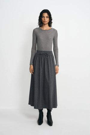 A model stands against a light gray studio background wearing a gray long-sleeve knit top and a matching gray midi skirt with a drawstring waist, paired with black ankle boots.