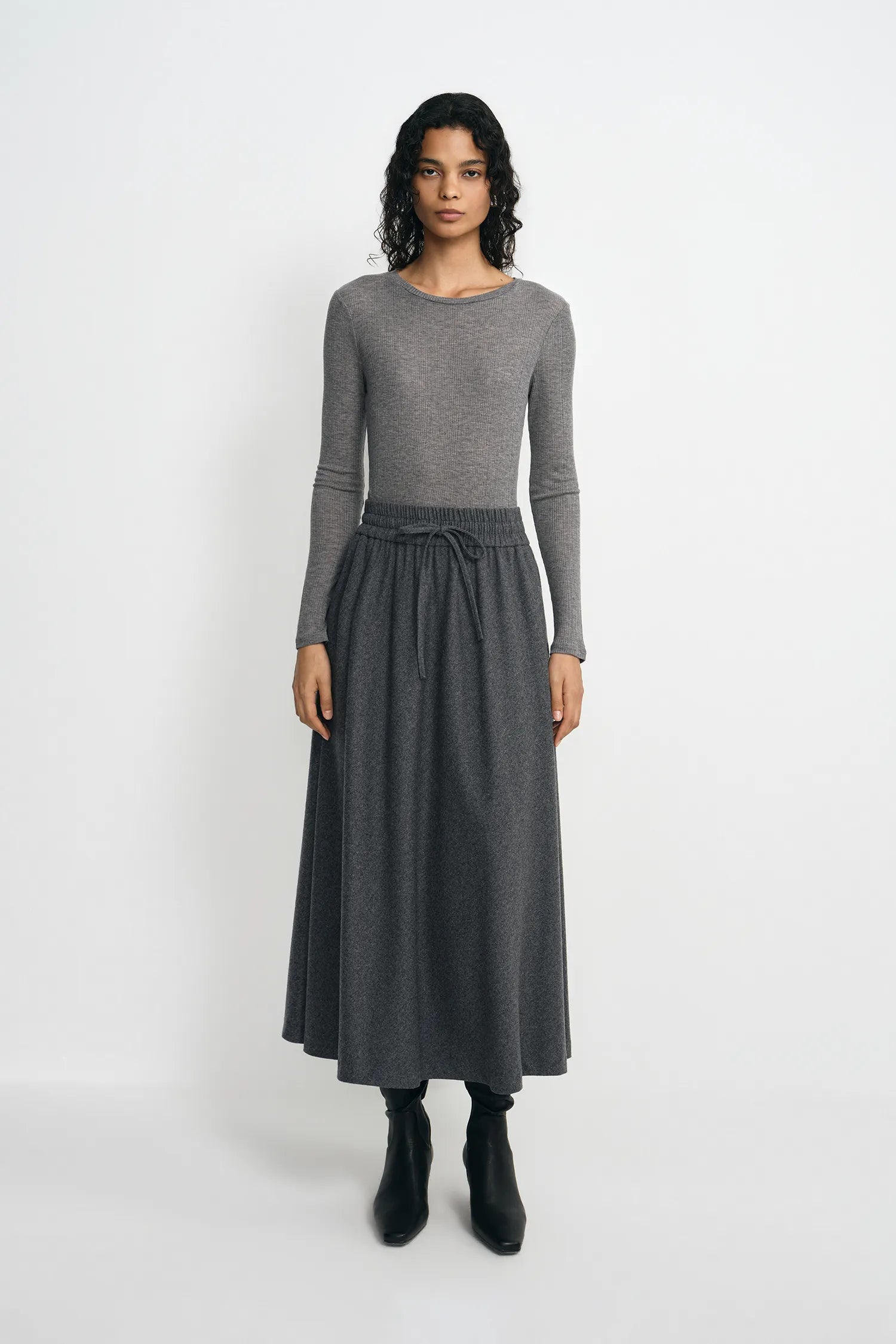 A model stands against a light gray studio background wearing a gray long-sleeve knit top and a matching gray midi skirt with a drawstring waist, paired with black ankle boots.