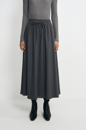 A model wearing a dark gray midi skirt with an elastic drawstring waist, paired with a gray long-sleeve top and black ankle boots, against a plain light gray background.