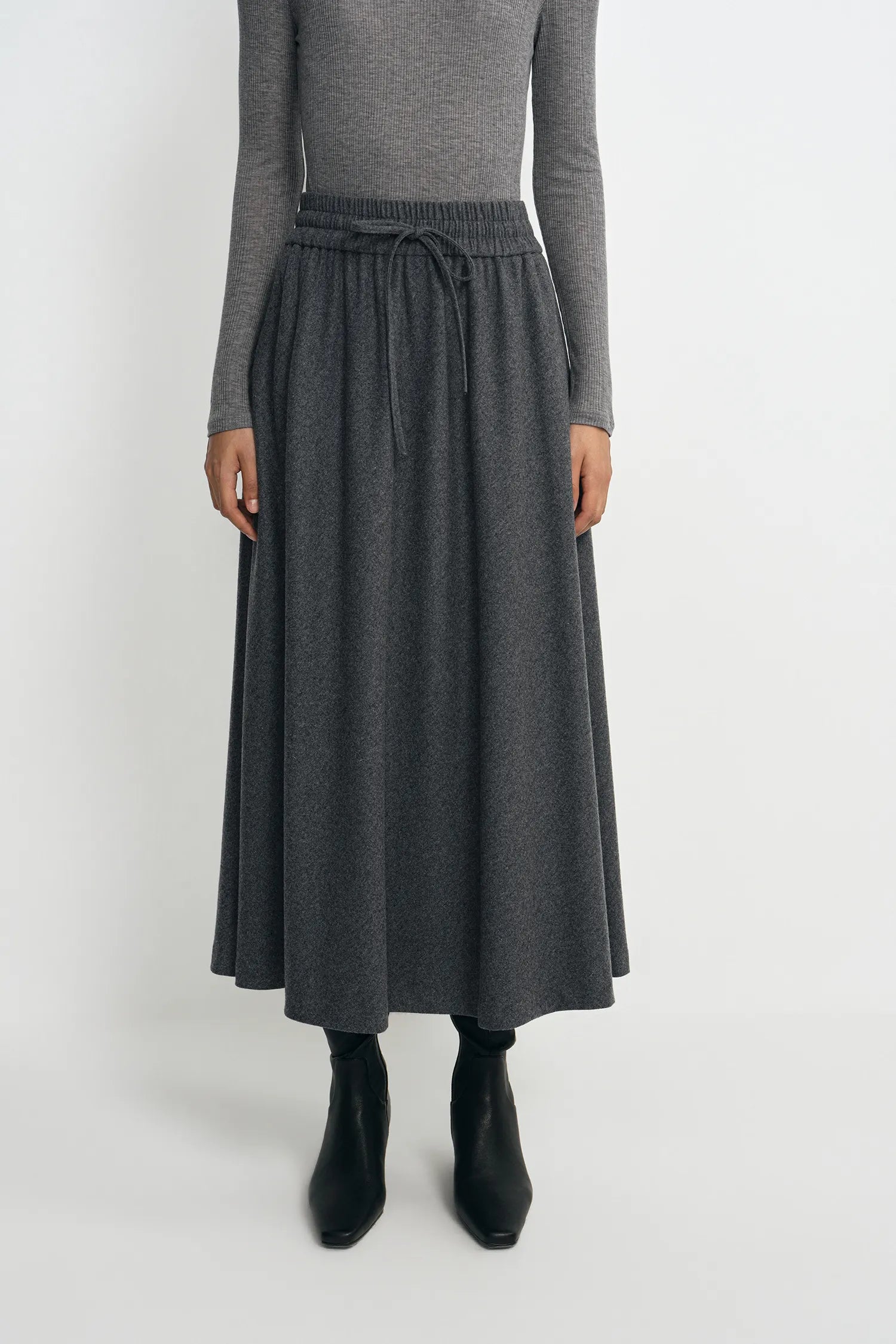 A model wearing a dark gray midi skirt with an elastic drawstring waist, paired with a gray long-sleeve top and black ankle boots, against a plain light gray background.