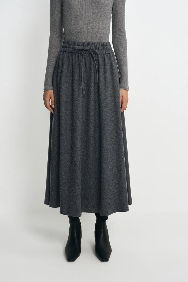 A model wearing a dark gray midi skirt with an elastic drawstring waist, paired with a gray long-sleeve top and black ankle boots, against a plain light gray background.