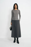 A model stands against a plain light gray studio background wearing a fitted long-sleeve gray top and a matching dark gray midi skirt, with black ankle boots and a small black handbag.