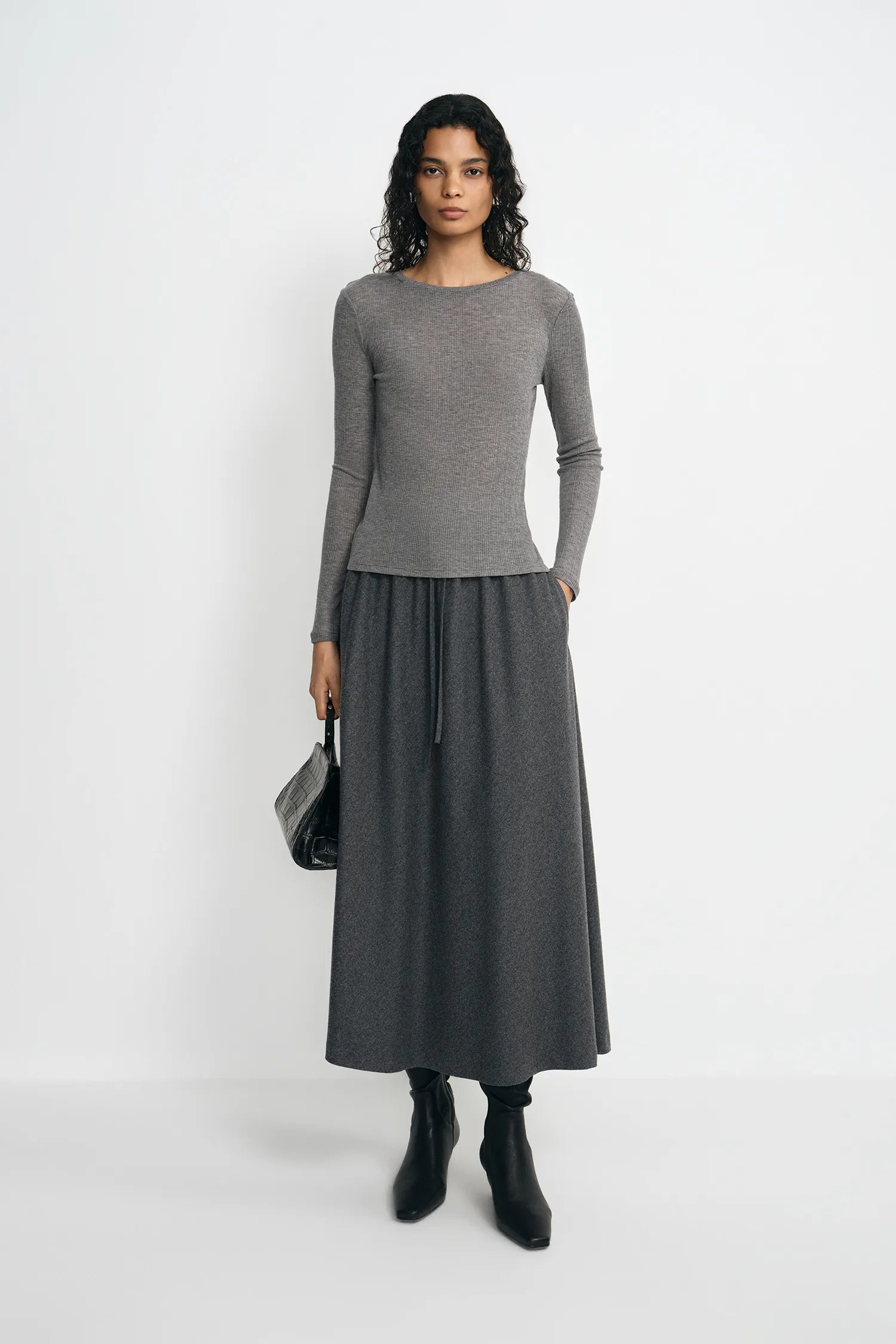 A model stands against a plain light gray studio background wearing a fitted long-sleeve gray top and a matching dark gray midi skirt, with black ankle boots and a small black handbag.