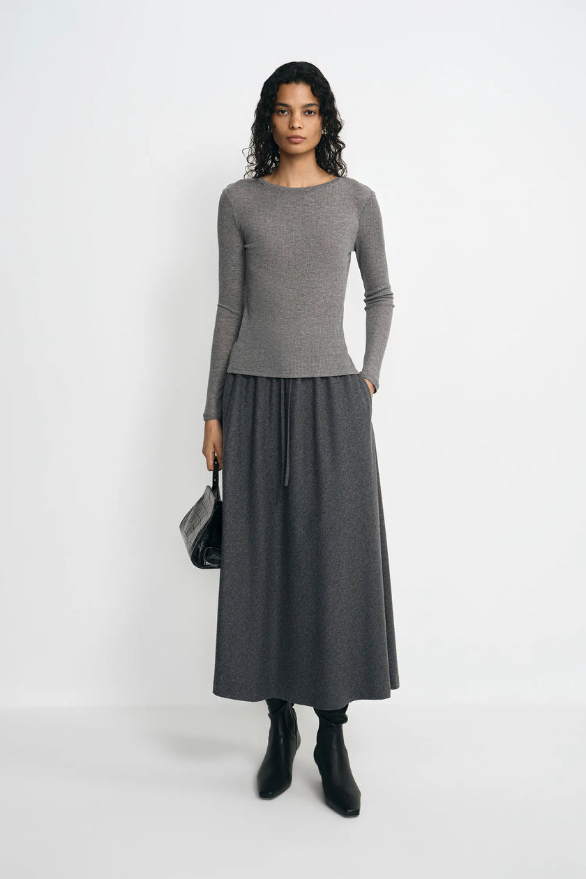A model stands against a plain light gray studio background wearing a fitted long-sleeve gray top and a matching dark gray midi skirt, with black ankle boots and a small black handbag.