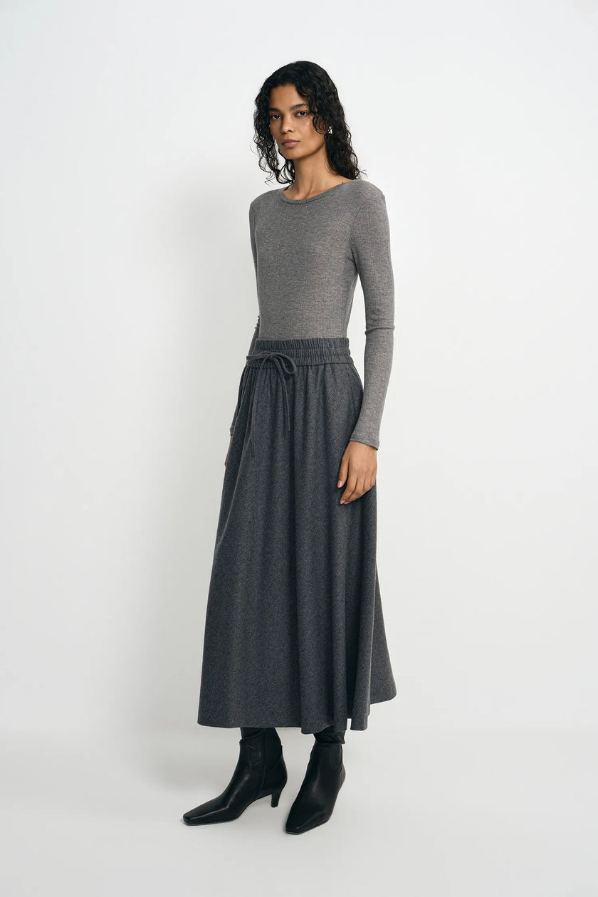 A woman modeling a gray long-sleeve knit top paired with a dark gray high-waisted midi skirt featuring a drawstring waist, styled with black ankle boots against a light gray studio background.