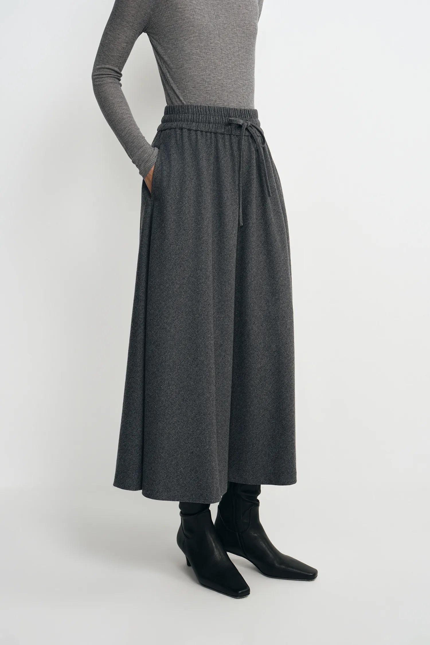 A model wearing a dark gray wide-leg midi skirt with an elastic waistband and drawstring, styled with a gray long-sleeve top and black ankle boots against a light gray studio background.