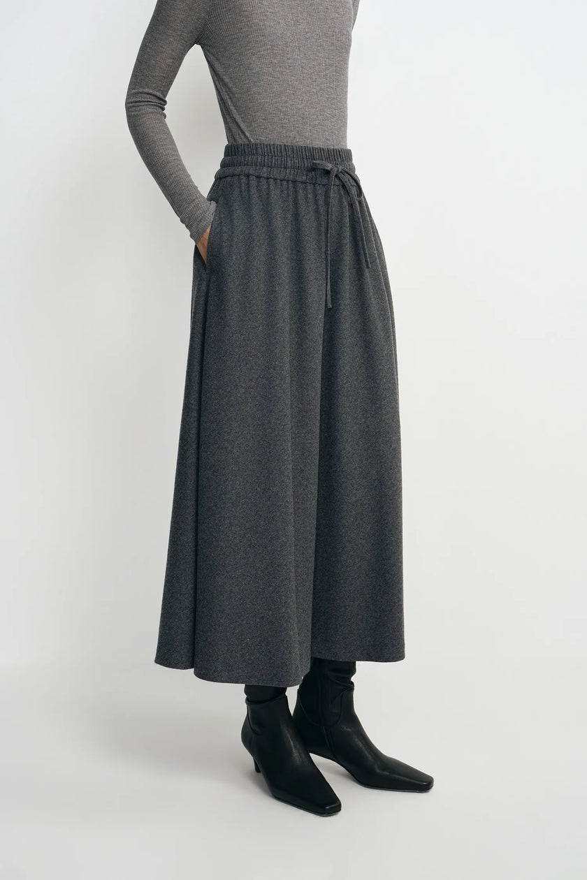 A model wearing a dark gray wide-leg midi skirt with an elastic waistband and drawstring, styled with a gray long-sleeve top and black ankle boots against a light gray studio background.