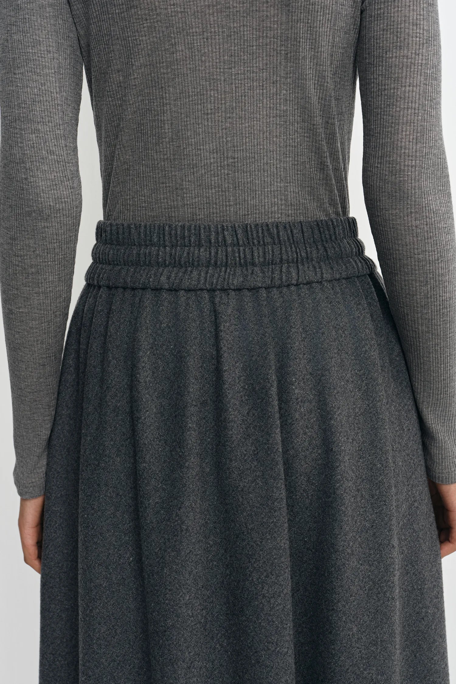 Close-up of a person wearing a dark gray ribbed knit long-sleeve top and a matching dark gray pleated skirt with an elastic waistband.