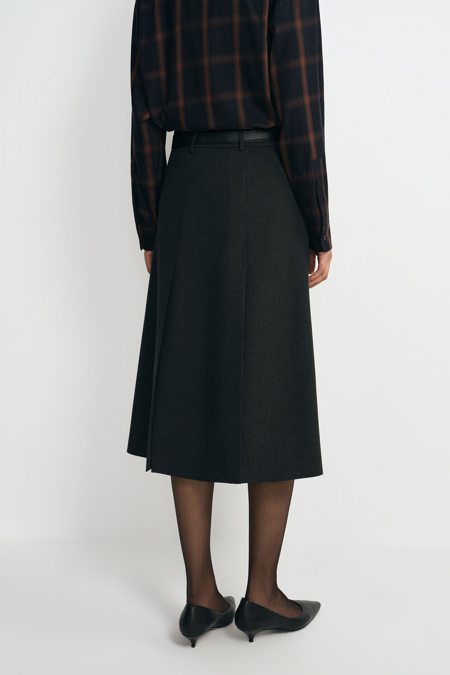 A model wearing a charcoal A-line skirt with a vented back pleat, pairing a plaid button-up shirt, back view.
