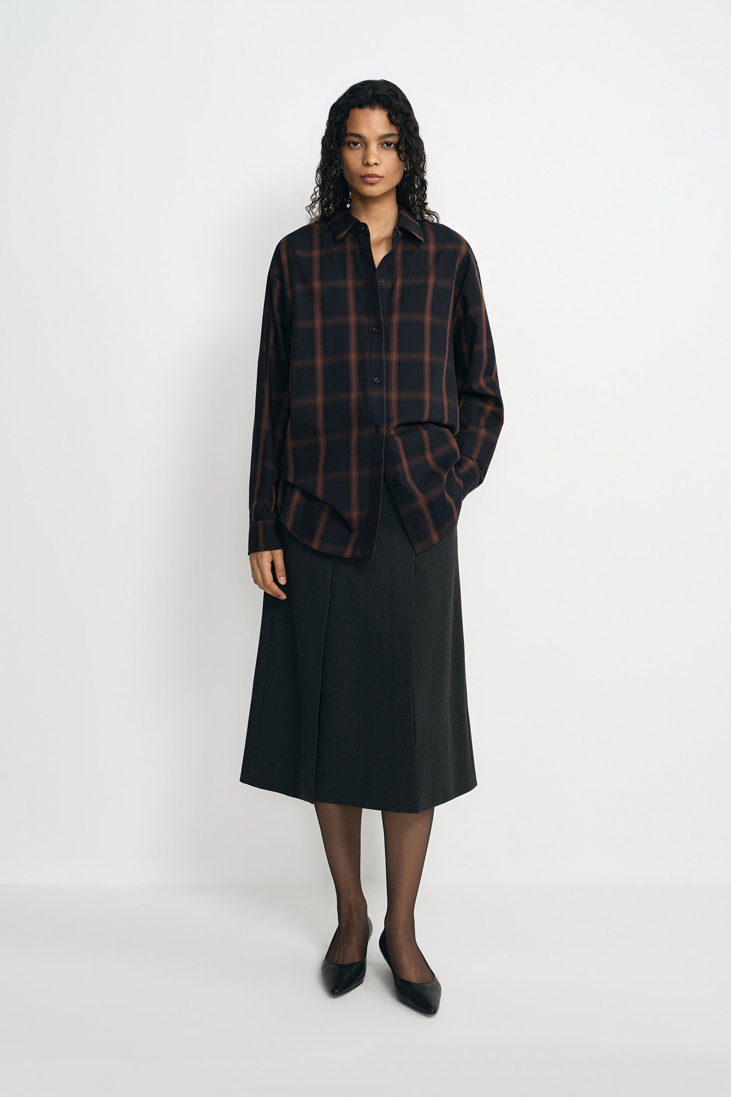 A model wearing a charcoal A-line skirt with a vented front detail, pairing a plaid button-up shirt, facing the camera.