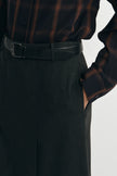 Close-up of a person wearing dark charcoal gray pleated trousers, a black leather belt with a silver buckle, and a dark brown and black plaid button-down shirt.