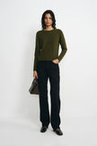 A model stands against a light gray studio background wearing an olive green long-sleeve knit top, black wide-leg trousers, black shoes, and holding a dark brown handbag.