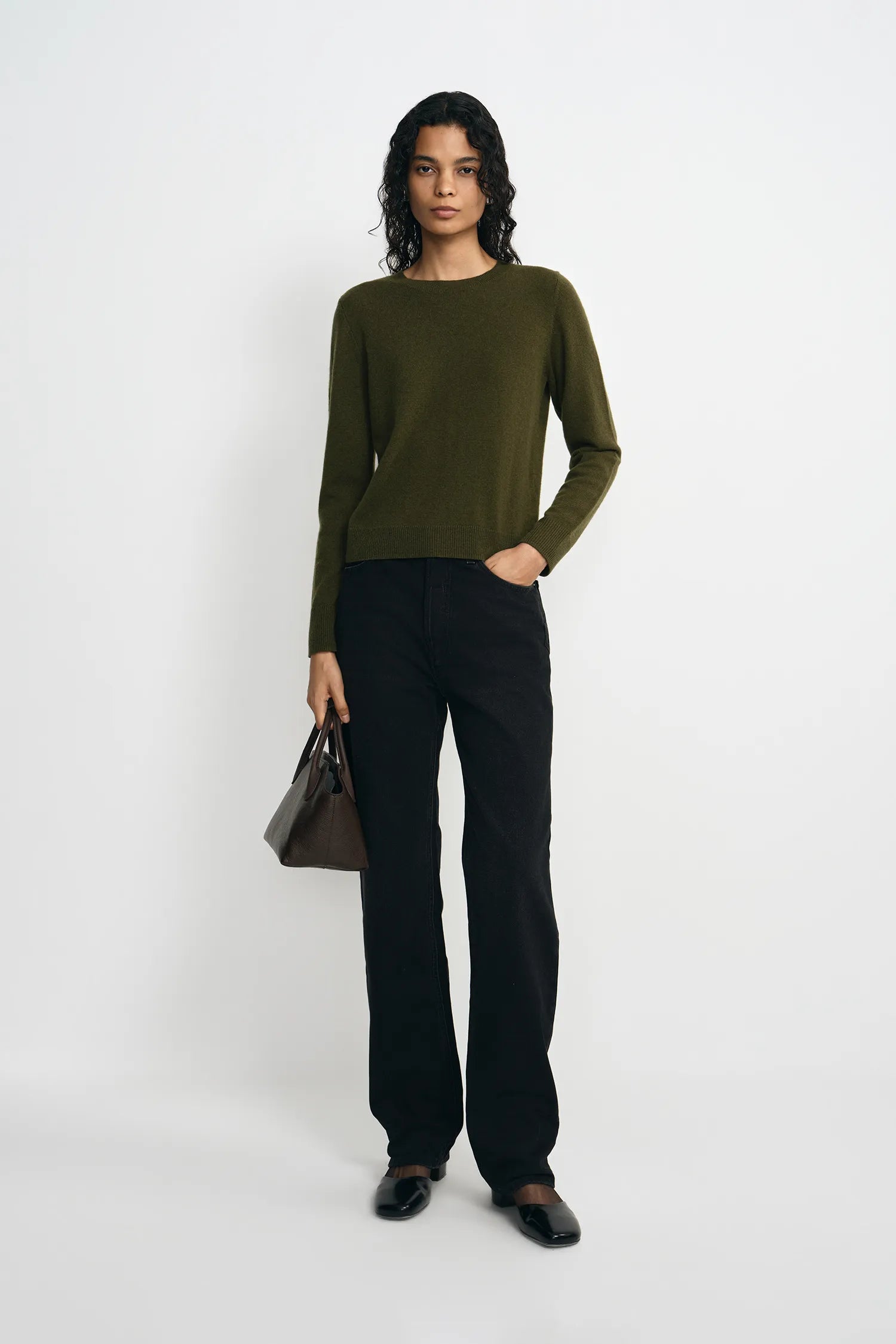 A model stands against a light gray studio background wearing an olive green long-sleeve knit top, black wide-leg trousers, black shoes, and holding a dark brown handbag.