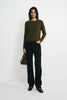 A model stands against a light gray studio background wearing an olive green long-sleeve knit top, black wide-leg trousers, black shoes, and holding a dark brown handbag.