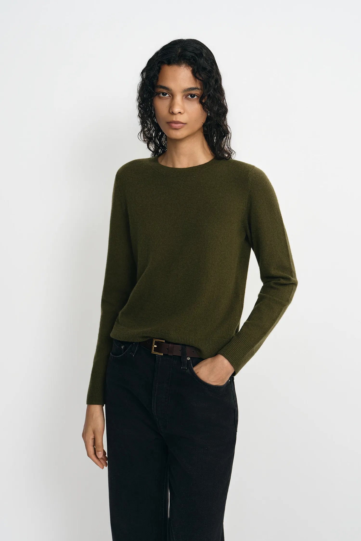 A model wearing an olive green long-sleeve knit sweater and black jeans, standing against a plain white studio background.