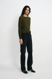 A model stands against a plain light gray studio background wearing an olive green long-sleeve knit sweater, black straight-leg jeans, and black shoes.