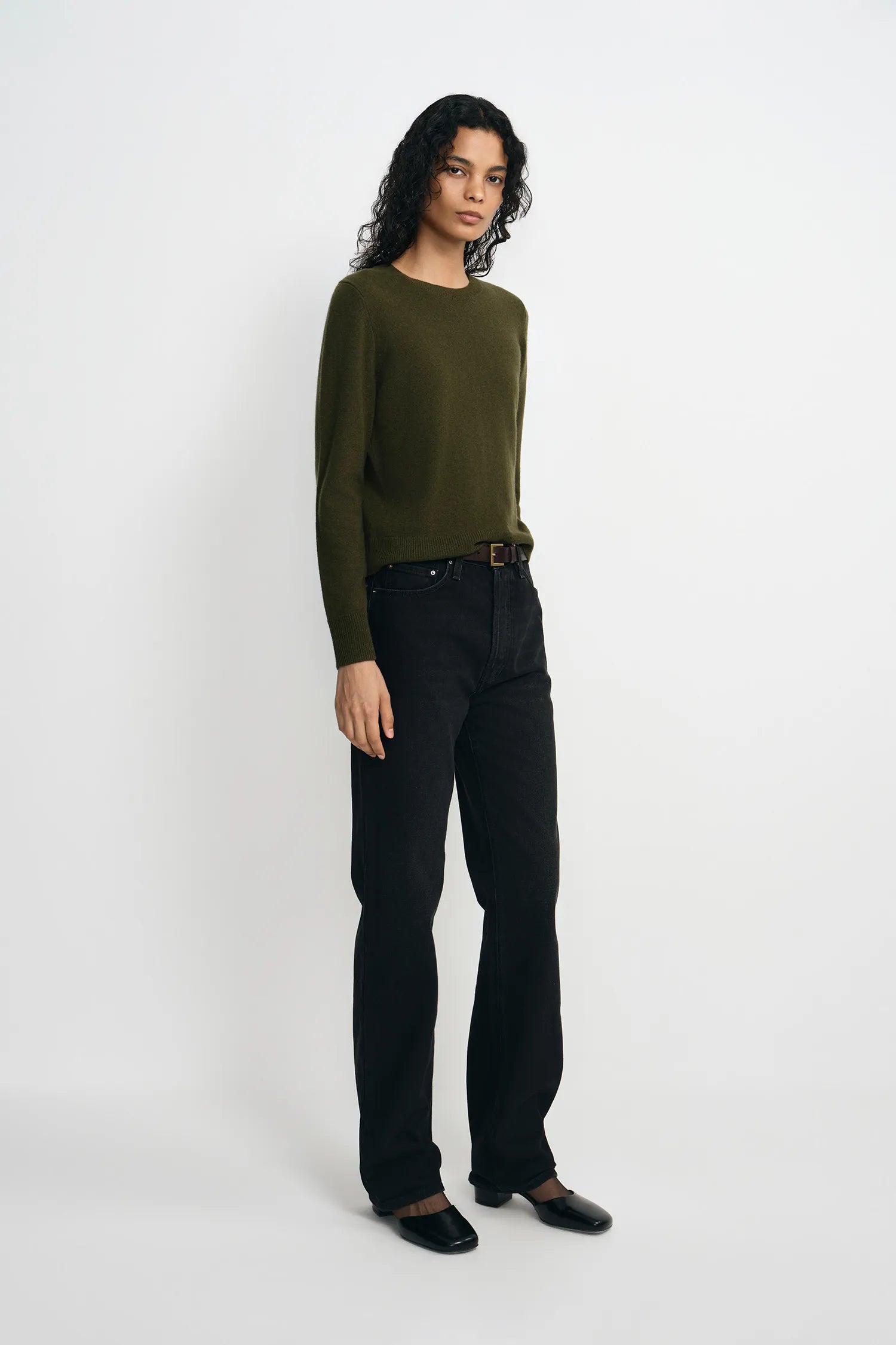 A model stands against a plain light gray studio background wearing an olive green long-sleeve knit sweater, black straight-leg jeans, and black shoes.