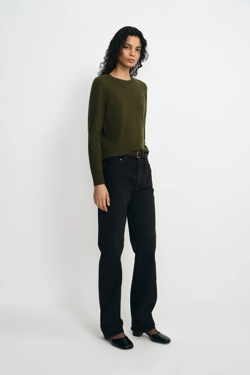 A model stands against a plain light gray studio background wearing an olive green long-sleeve knit sweater, black straight-leg jeans, and black shoes.