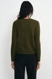 A woman viewed from the back wearing an olive green long-sleeve knit sweater and black pants against a plain light gray studio background.