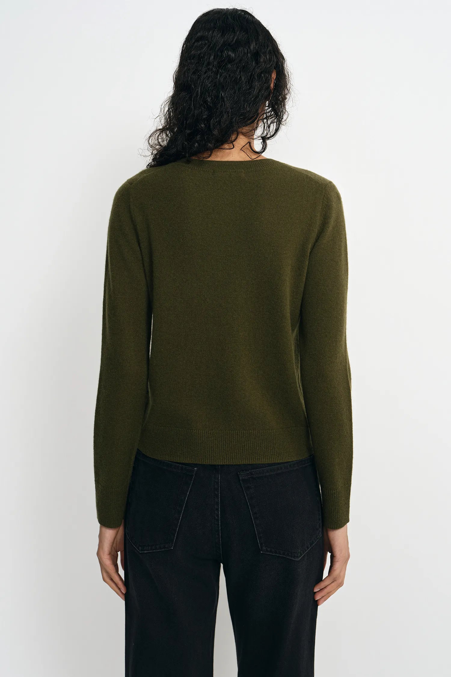 A woman viewed from the back wearing an olive green long-sleeve knit sweater and black pants against a plain light gray studio background.