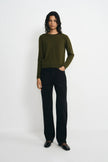 A model stands against a light gray studio background wearing an olive green long-sleeve knit sweater, black wide-leg trousers, and black shoes.