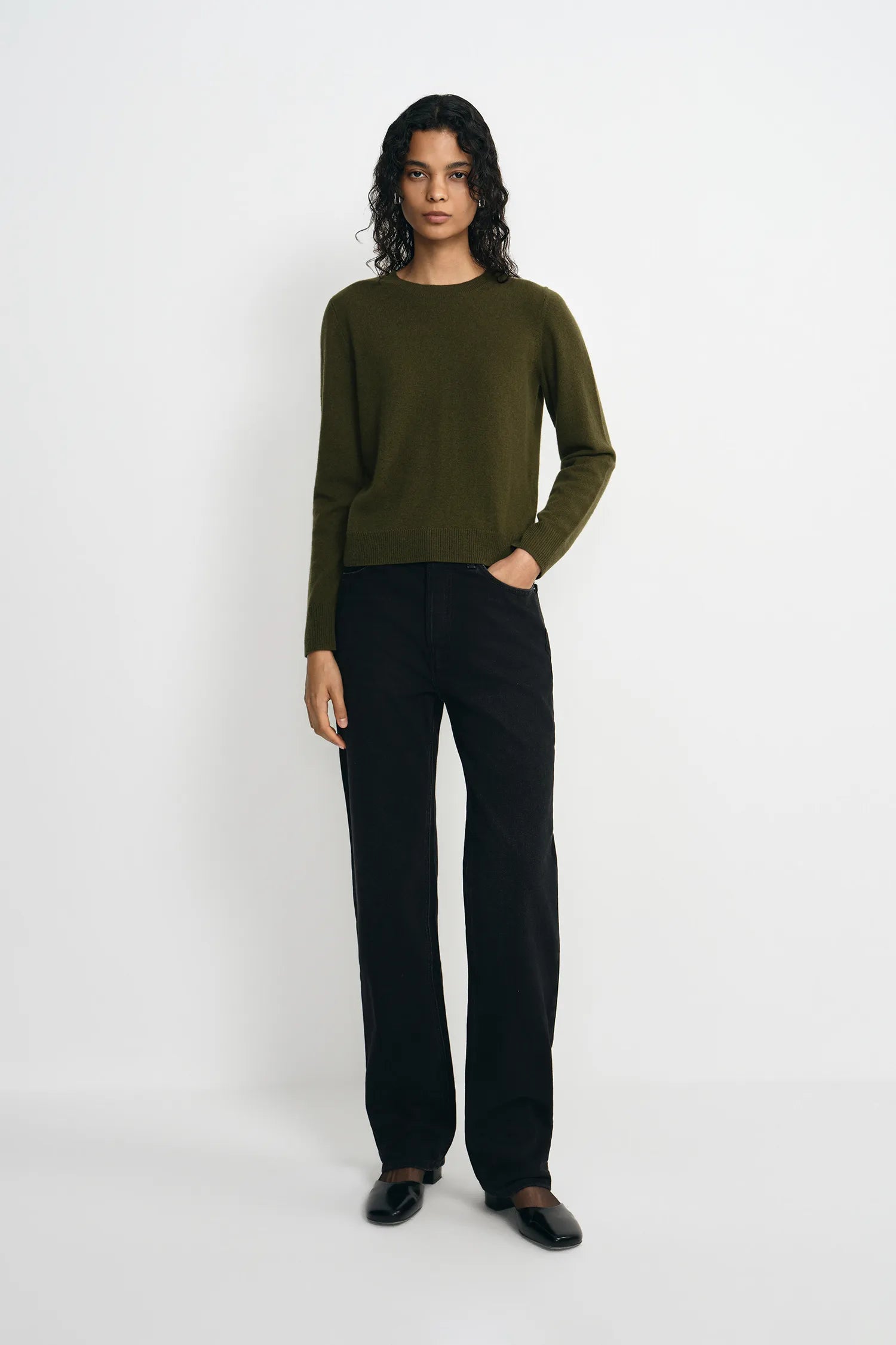 A model stands against a light gray studio background wearing an olive green long-sleeve knit sweater, black wide-leg trousers, and black shoes.