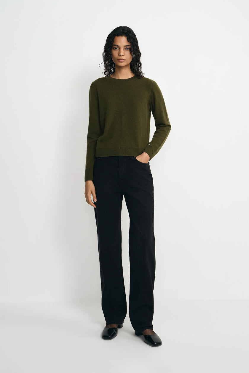 A model stands against a light gray studio background wearing an olive green long-sleeve knit sweater, black wide-leg trousers, and black shoes.