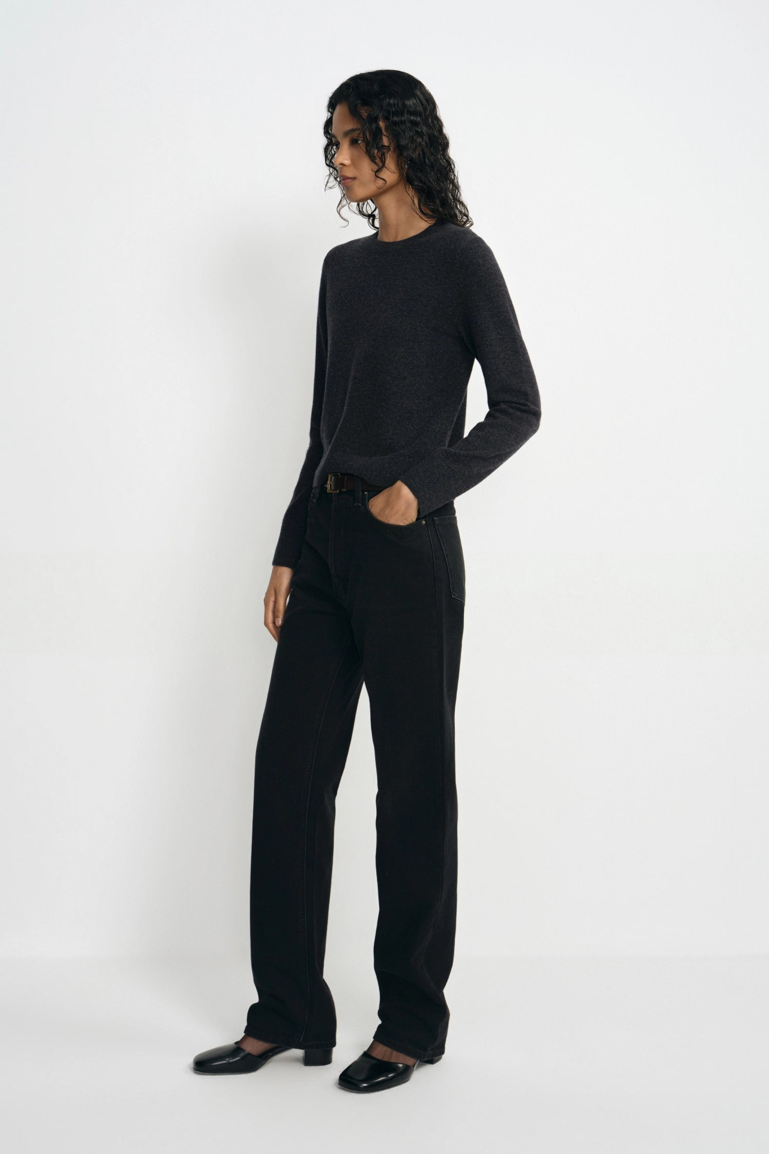 A model wearing a dark charcoal gray long-sleeve knit top and black wide-leg trousers, standing in profile against a clean white studio background.