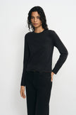 A model wearing a dark charcoal gray long-sleeve knit sweater and black jeans, standing against a light gray studio background.