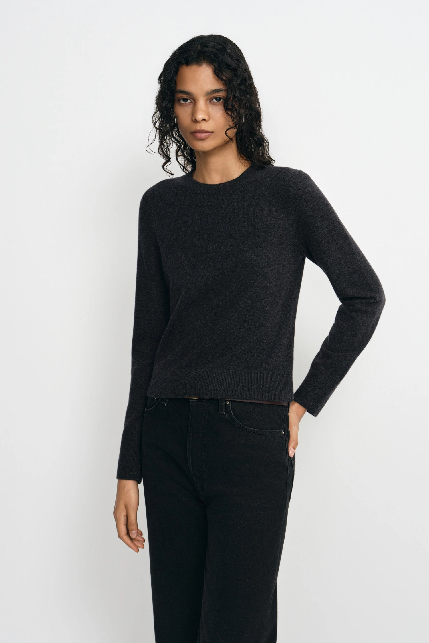 A model wearing a dark charcoal gray long-sleeve knit sweater and black jeans, standing against a light gray studio background.