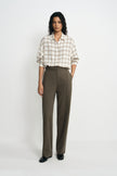 A model stands against a light gray studio background wearing a cream and tan plaid button-down shirt tucked into high-waisted, wide-leg olive brown trousers, paired with black shoes.