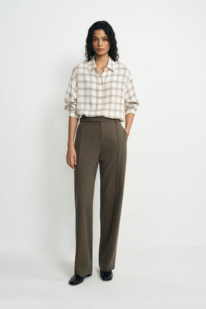 A model stands against a light gray studio background wearing a cream and tan plaid button-down shirt tucked into high-waisted, wide-leg olive brown trousers, paired with black shoes.