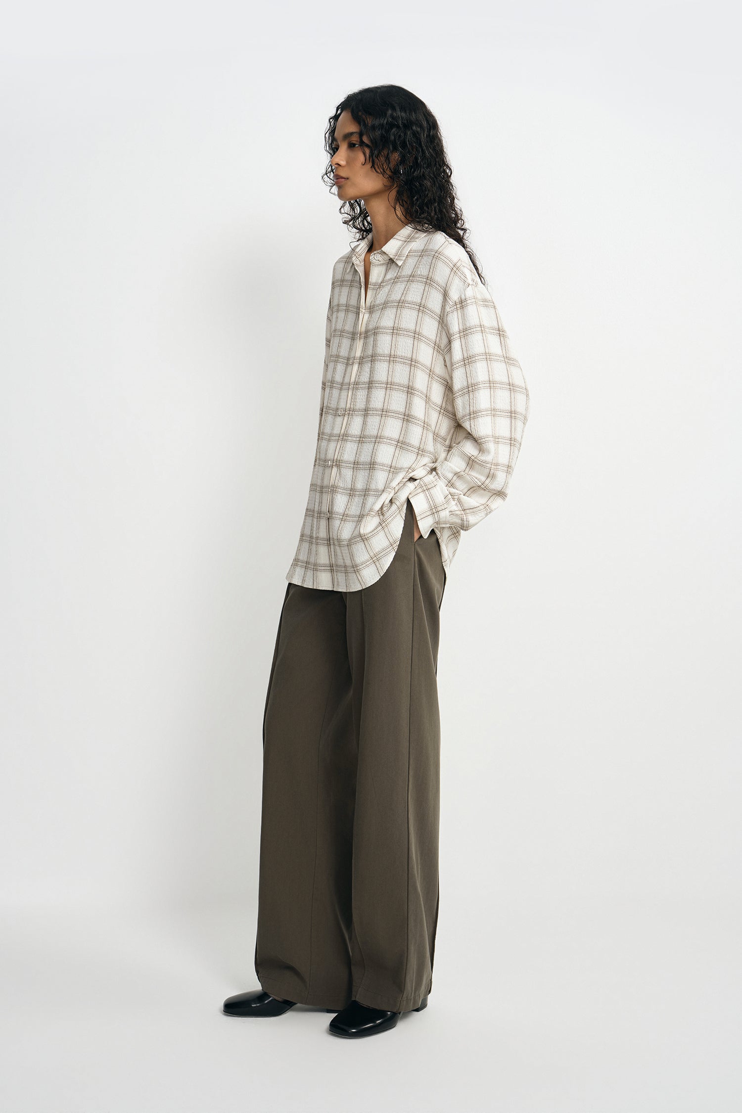 A model stands in profile wearing a cream and beige plaid button-down shirt with wide-leg olive brown trousers and black shoes, set against a plain light gray studio background.