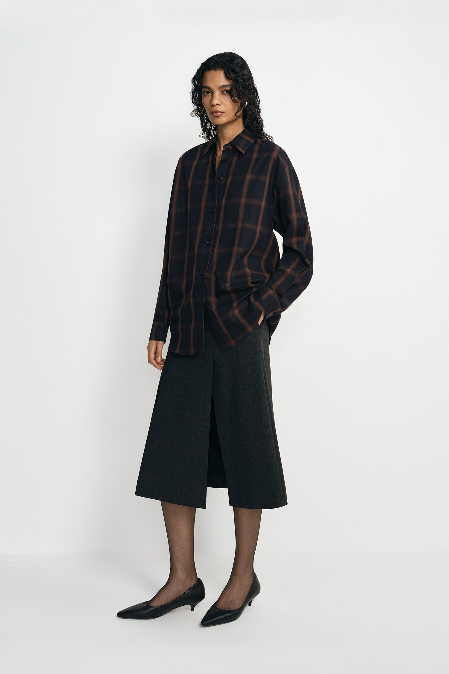 A model wearing a plaid button-up shirt, pairing a charcoal A-line midi skirt, turned slightly to the side.