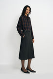 A model wearing a plaid button-up shirt, pairing a charcoal pleated midi skirt with a belted waist, turned slightly to the side.