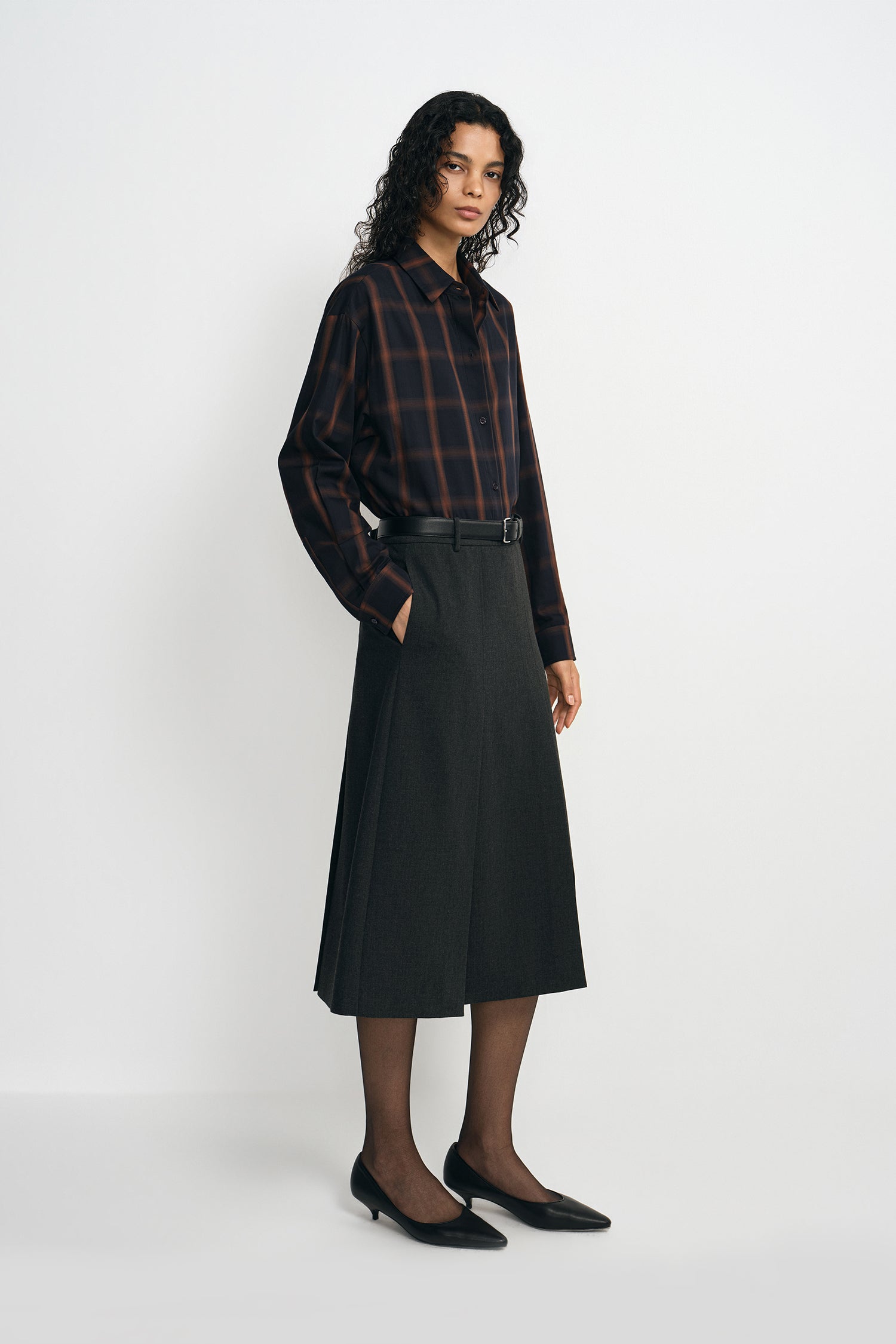 A model wearing a plaid button-up shirt, pairing a charcoal pleated midi skirt with a belted waist, turned slightly to the side.