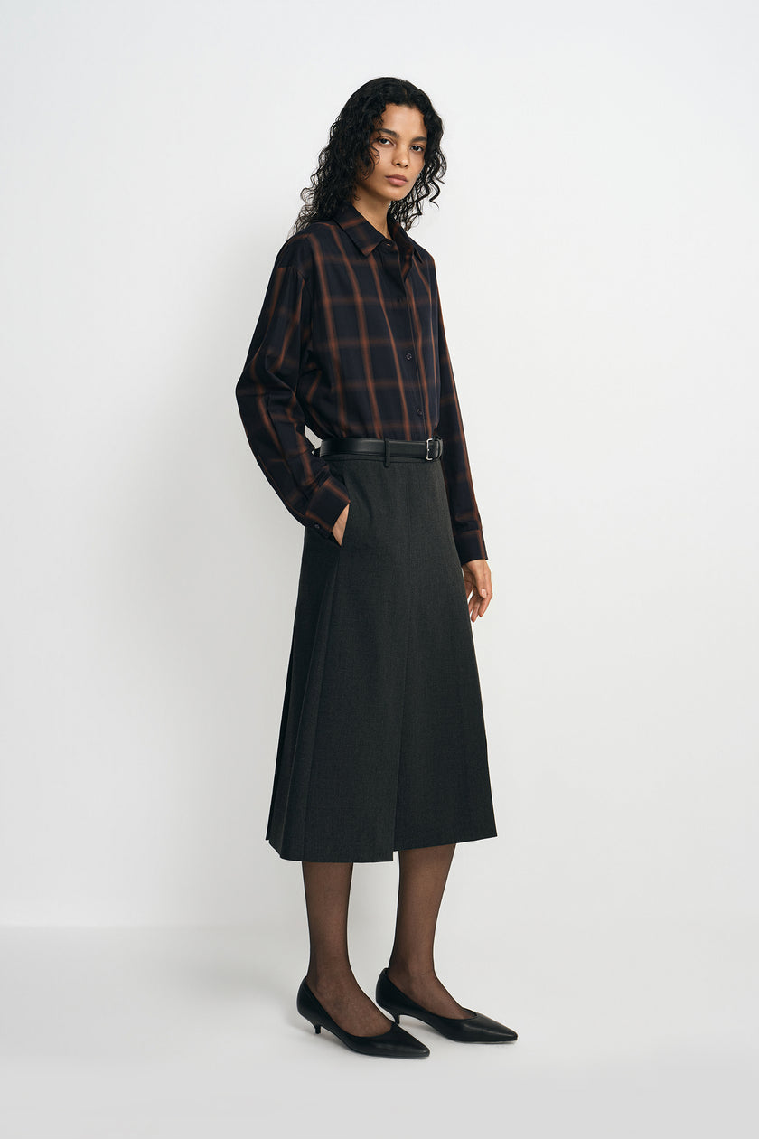 A model wearing a plaid button-up shirt, pairing a charcoal pleated midi skirt with a belted waist, turned slightly to the side.