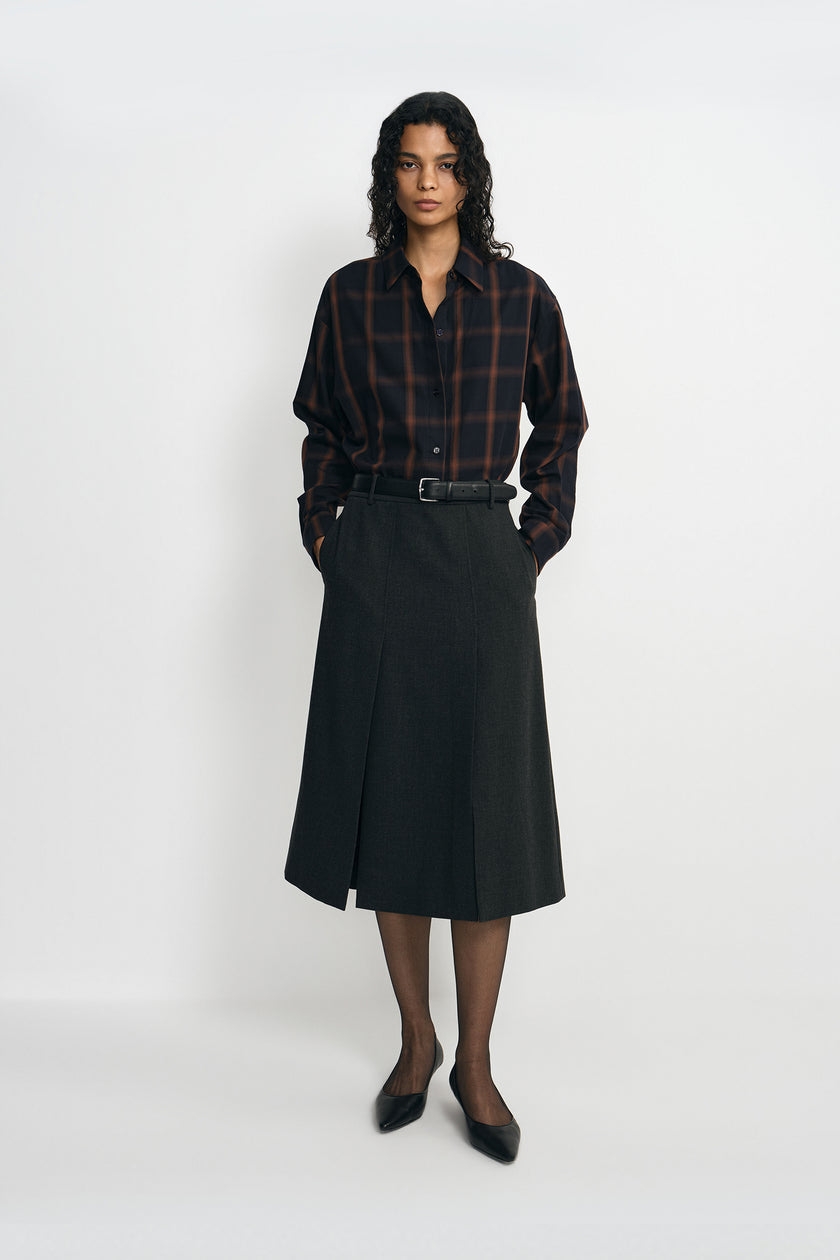A model wearing a plaid button-up shirt, pairing a charcoal pleated midi skirt with a belted waist.