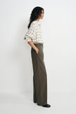 A woman stands in profile wearing a cream and tan plaid button-up shirt with rolled sleeves and dark olive wide-leg trousers, paired with black shoes, against a plain light gray studio background.