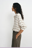A woman shown in side profile wearing a cream and brown plaid button-up shirt with rolled sleeves and dark olive pants, standing against a plain light gray studio background.
