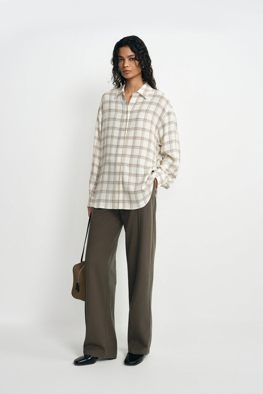 A model wearing a fluid plaid shirt, pairing forest wide-leg trousers, turned slightly to the side.
