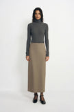 A model stands against a plain white studio background wearing a dark gray ribbed turtleneck sweater and a long taupe skirt, paired with black Mary Jane shoes.
