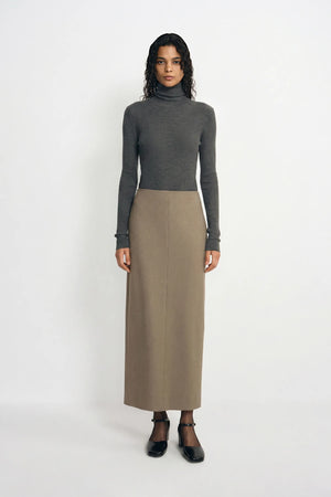 A model stands against a plain white studio background wearing a dark gray ribbed turtleneck sweater and a long taupe skirt, paired with black Mary Jane shoes.