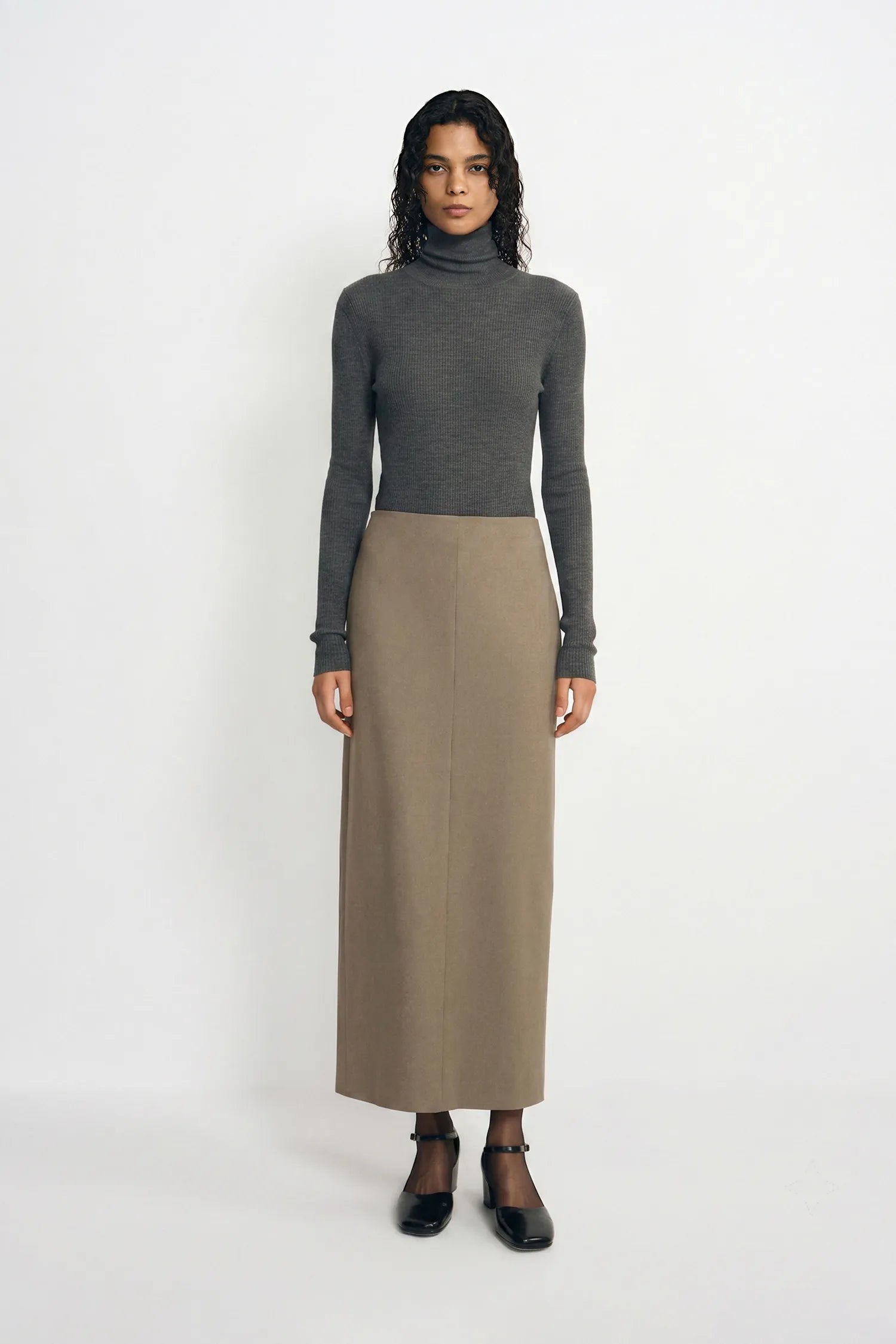 A model stands against a plain white studio background wearing a dark gray ribbed turtleneck sweater and a long taupe skirt, paired with black Mary Jane shoes.